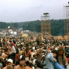 Tłum ludzi na festiwalu muzycznym, gdzie jest Woodstock. Widać scenę, wieże głośnikowe i namioty.