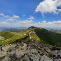 Zielone wzgórza Masywu Centralnego, pnące się ku błękitnemu niebu z białymi chmurami. Kręte ścieżki wiją się po zboczach, zapraszając do wędrówki.