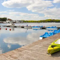 Słoneczny dzień nad jeziorem, gdzie miasto nad jeziorem zaprasza do wodnych przygód. Widać łódź żaglową, motorową i rzędy rowerów wodnych.
