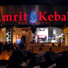 Jasny neon "Amrit Kebab" nad wejściem do restauracji. W tle widać wnętrze z pracującymi kucharzami i lodówką Coca-Coli. Na pierwszym planie stoją skutery z pudełkami na dowóz.