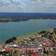 Tłum ludzi korzysta z uroków Jeziora Białego. Widać plażę, kąpielisko, budynki i las. Mnóstwo atrakcji czeka na turystów.