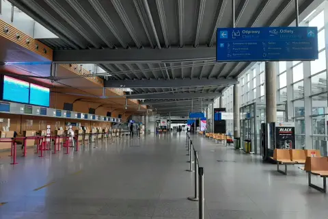 Pusta hala odlotów na lotnisku, z rzędami pustych stanowisk odprawy i miejscami do siedzenia. W tle widoczne tablice informacyjne, część poznania z portem lotniczym.