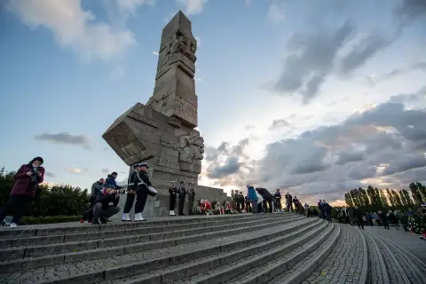 Uroczyste zwiedzanie Westerplatte. Monument, żołnierze w mundurach, wieńce i chmury na niebie tworzą podniosłą atmosferę.