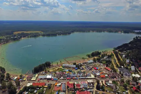 Tłum ludzi korzysta z uroków Jeziora Białego. Widać plażę, kąpielisko, budynki i las. Mnóstwo atrakcji czeka na turystów.