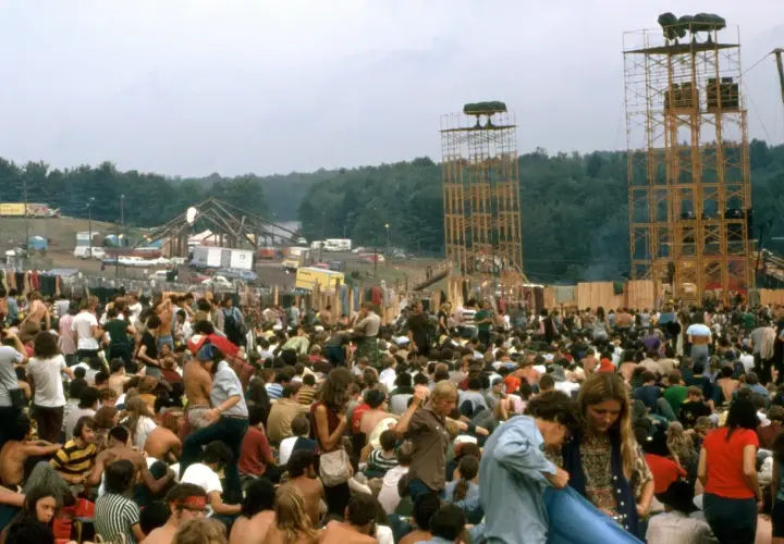 Tłum ludzi na festiwalu muzycznym, gdzie jest Woodstock. Widać scenę, wieże głośnikowe i namioty.