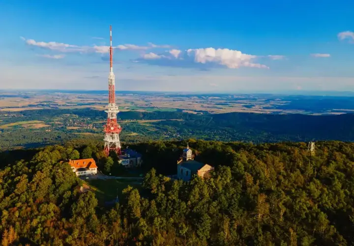 Wieża telekomunikacyjna na szczycie Ślęży, otoczona lasem i budynkami. Widok na rozległą dolinę.