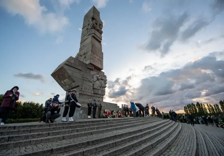 Uroczyste zwiedzanie Westerplatte. Monument, żołnierze w mundurach, wieńce i chmury na niebie tworzą podniosłą atmosferę.