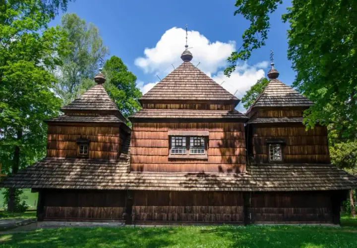 Drewniana cerkiew z charakterystycznymi kopułami, otoczona zielenią. W oddali widać krajobraz, a na pierwszym planie trawa. To miejsce, gdzie czas się zatrzymał, jak w dawnych lutowiskach.