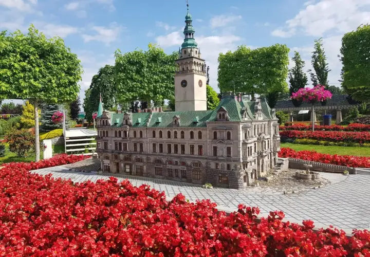 Miniatura ratusza z wieżą zegarową, otoczona czerwonymi kwiatami, w parku miniatur w Kotlinie Kłodzkiej.