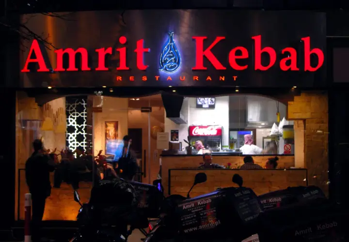 Jasny neon "Amrit Kebab" nad wejściem do restauracji. W tle widać wnętrze z pracującymi kucharzami i lodówką Coca-Coli. Na pierwszym planie stoją skutery z pudełkami na dowóz.