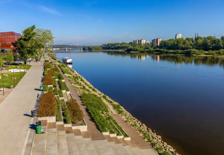 Nowoczesny boulevard nad rzeką z zielenią, nowoczesnym budynkiem i mostem w oddali.