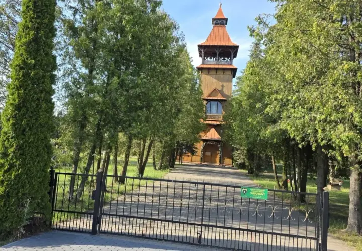 Drewniana wieża kościelna w Długosiodle, otoczona drzewami, za żelazną bramą.
