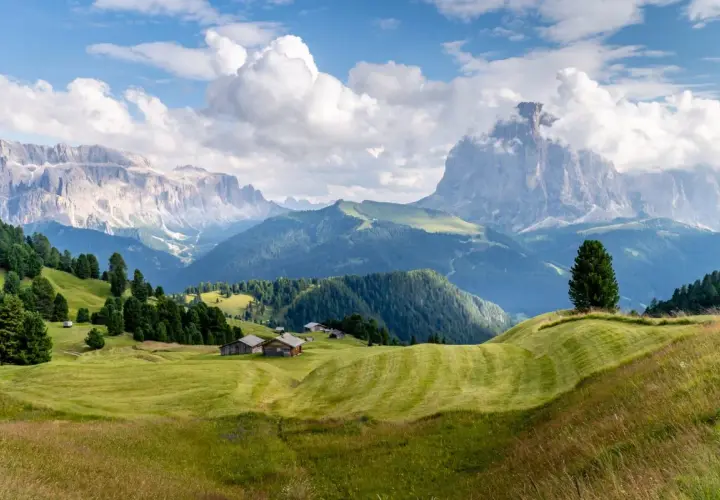 Zielone wzgórza i drewniane chatki w Val Gardena, z majestatycznymi Dolomitami w tle.