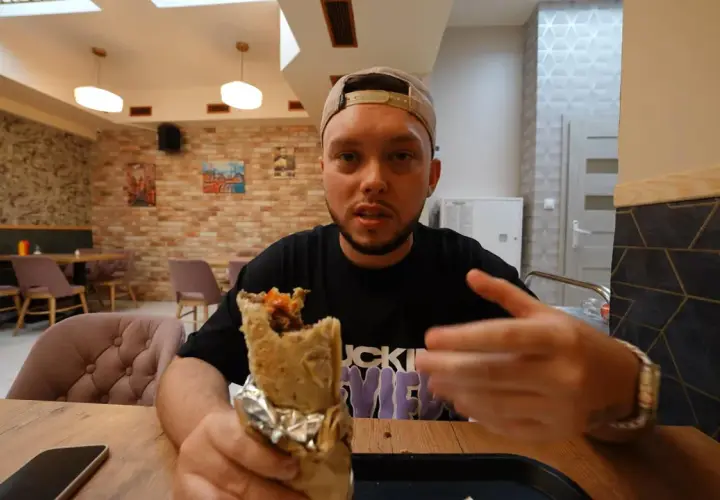 Mężczyzna w czapce z daszkiem trzyma w ręku dużego kebaba, wyglądając jak prawdziwy lider kebab. W tle widać wnętrze restauracji.