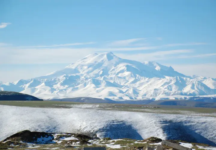 Majestatyczny, ośnieżony najwyższy szczyt Kaukazu, Elbrus, góruje nad pokrytymi śniegiem wzgórzami i zielonymi dolinami.