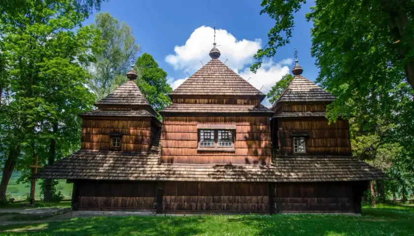 Drewniana cerkiew z charakterystycznymi kopułami, otoczona zielenią. W oddali widać krajobraz, a na pierwszym planie trawa. To miejsce, gdzie czas się zatrzymał, jak w dawnych lutowiskach.