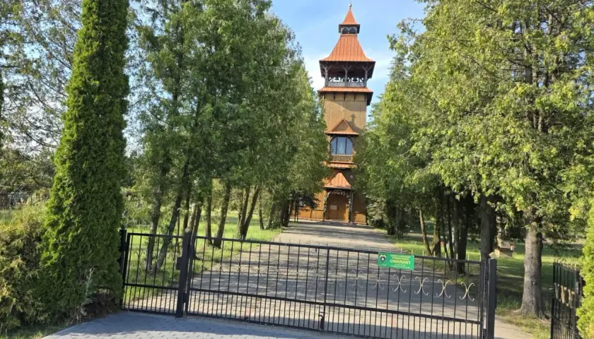 Drewniana wieża kościelna w Długosiodle, otoczona drzewami, za żelazną bramą.