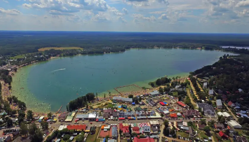 Tłum ludzi korzysta z uroków Jeziora Białego. Widać plażę, kąpielisko, budynki i las. Mnóstwo atrakcji czeka na turystów.