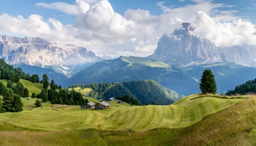 Zielone wzgórza i drewniane chatki w Val Gardena, z majestatycznymi Dolomitami w tle.