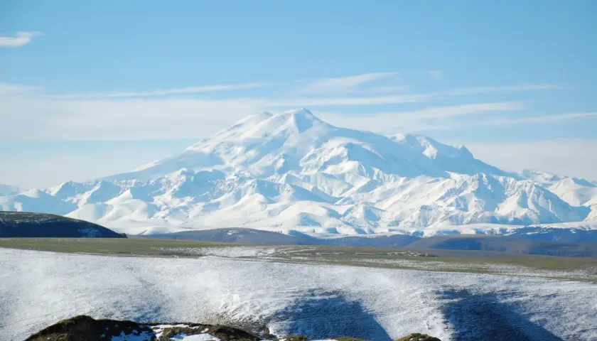 Majestatyczny, ośnieżony najwyższy szczyt Kaukazu, Elbrus, góruje nad pokrytymi śniegiem wzgórzami i zielonymi dolinami.