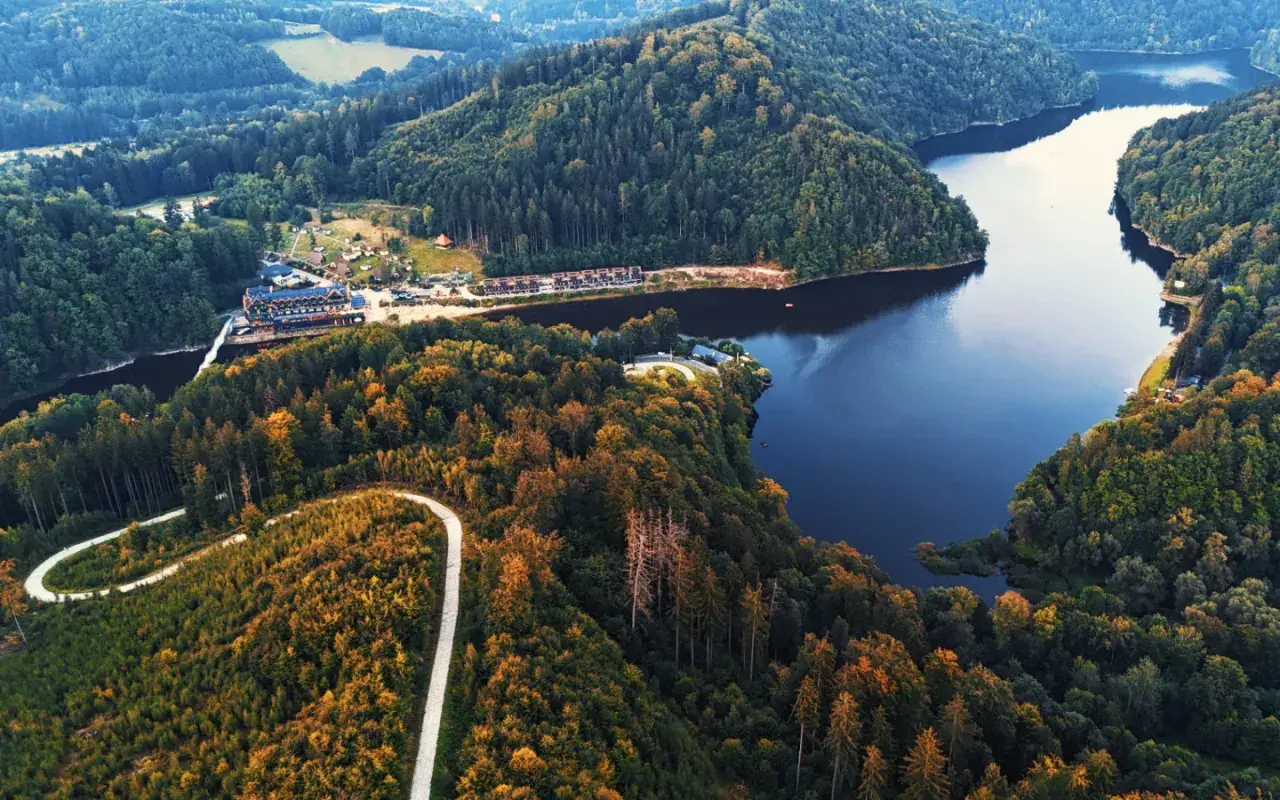 Malownicze jezioro Bystrzyckie otoczone lasami, z krętą drogą wijącą się po zboczu i hotelem nad wodą.