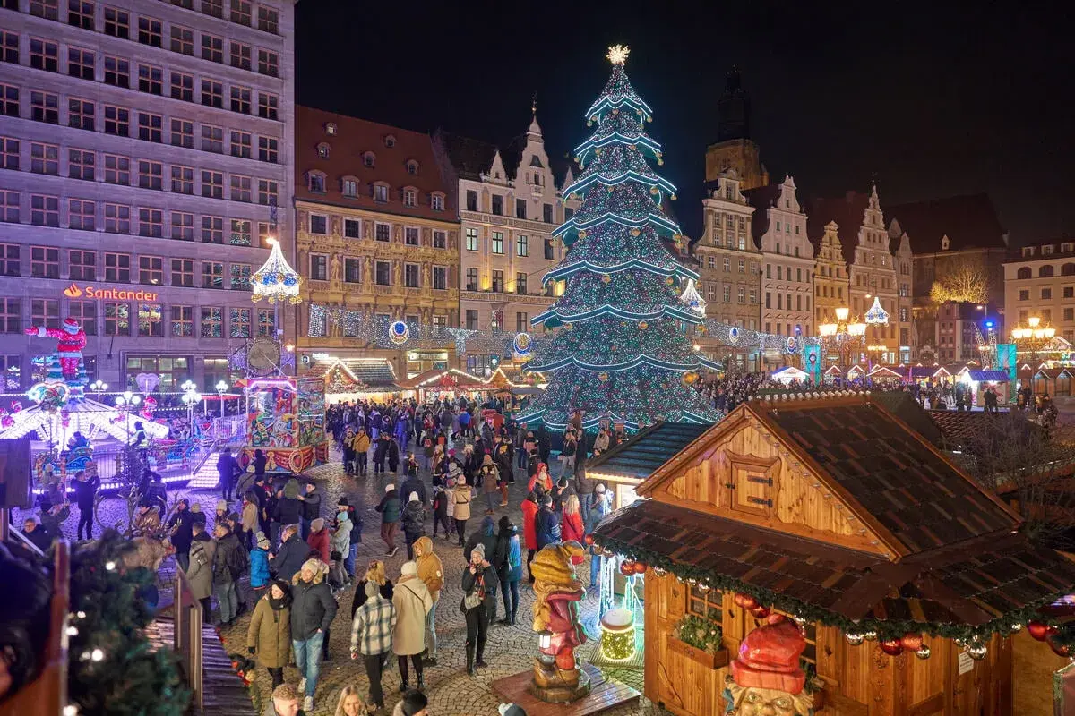 Tłoczny jarmark we Wrocławiu nocą. Rozświetlona choinka, kramy i tłumy ludzi tworzą magiczną atmosferę.