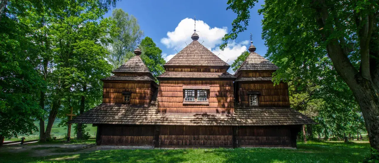 Drewniana cerkiew z charakterystycznymi kopułami, otoczona zielenią. W oddali widać krajobraz, a na pierwszym planie trawa. To miejsce, gdzie czas się zatrzymał, jak w dawnych lutowiskach.