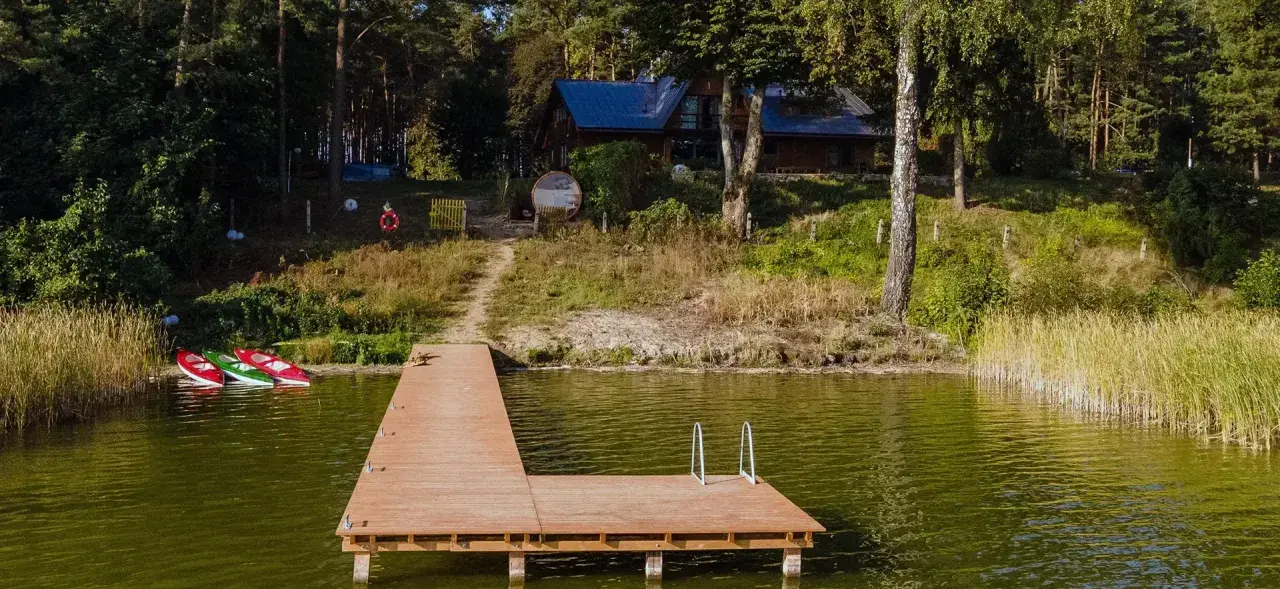 Drewniany pomost nad jeziorem Kałębie, z trzema kajakami przy brzegu. W tle las i dom.