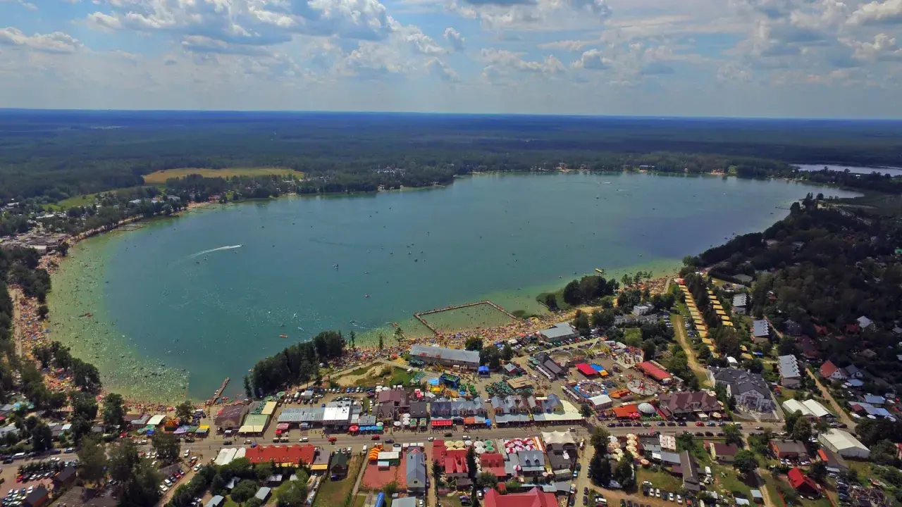 Tłum ludzi korzysta z uroków Jeziora Białego. Widać plażę, kąpielisko, budynki i las. Mnóstwo atrakcji czeka na turystów.