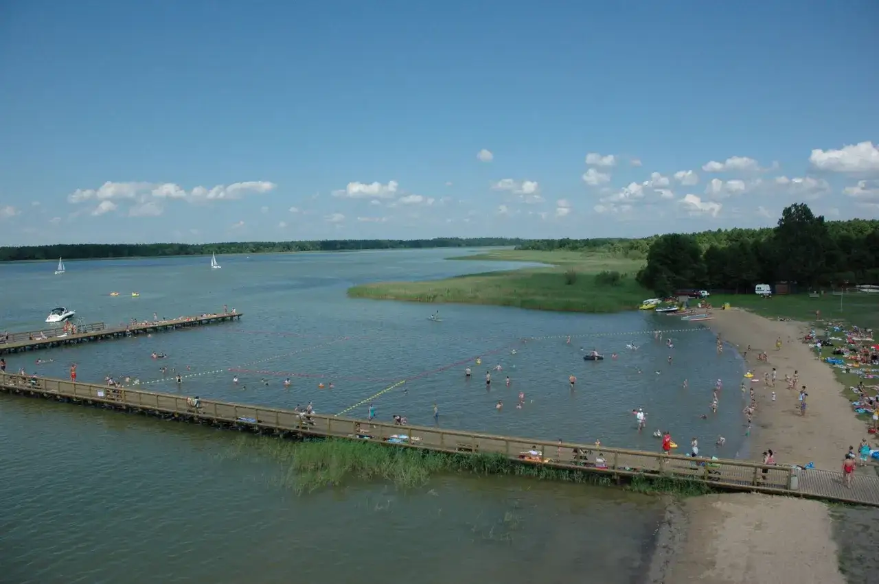 Letnie popołudnie nad jeziorem. Ludzie kąpią się, opalają na plaży i spacerują po drewnianych pomostach. W oddali widać żaglówki i łodzie. To idealne miejsce dla każdego miasto nad jeziorem roś.
