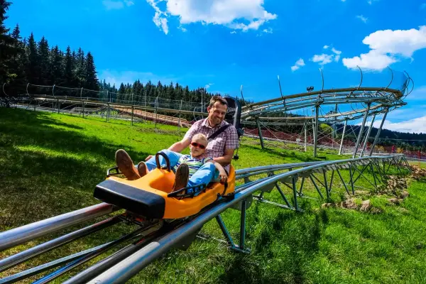 Ojciec i syn z uśmiechem zjeżdżają na sankach po torze zjazdowym. W tle widać czarną górną część konstrukcji toru.